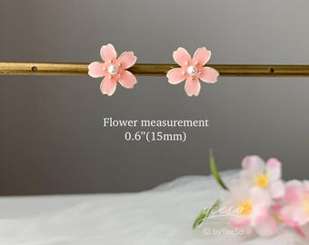 May include: A pair of pink flower stud earrings with a pearl center. The flower measures 0.6 inches (15mm) across. The earrings are set against a neutral background. The image also includes the text "Flower measurement 0.6"(15mm)".