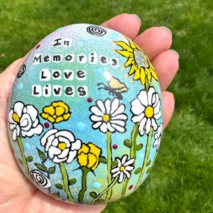 May include: Hand-painted stone with a floral design and the words "In Memories Love Lives." The artwork features white daisies, yellow flowers, a sunflower, and a butterfly against a blue and green background. The stone is held in a hand.
