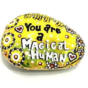May include: A smooth, oval-shaped river rock painted in bright yellow and green with a floral pattern. The text "You are a MAGICAL HUMAN" is written in black and white lettering across the center, accompanied by small painted heart icons.