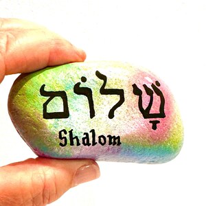 May include: A smooth, oval-shaped river stone painted with a vibrant, multi-colored pastel gradient of pink, yellow, green, and blue. The Hebrew word Shalom is written in bold black lettering, with the English word Shalom printed directly below it.