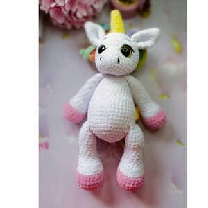 Può includere: Un giocattolo di unicorno bianco all'uncinetto con zoccoli rosa e un corno giallo. L'unicorno ha occhi neri e un'espressione amichevole.