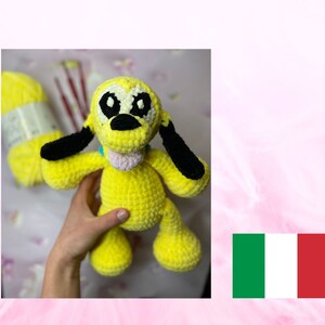 Può includere: Un cane all'uncinetto giallo fatto a mano con orecchie e occhi neri, una lingua rosa e un collare blu. Il peluche è tenuto in mano da una persona. Un gomitolo di lana gialla e ferri da maglia sono sullo sfondo.