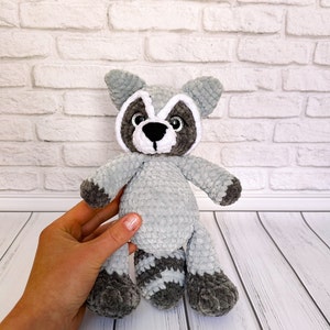 Può includere: Un peluche di procione all'uncinetto fatto a mano nei toni del grigio e del nero. L'animale di peluche ha una maschera distintiva intorno agli occhi, una coda a strisce e piccole orecchie. Il giocattolo morbido è tenuto in mano contro un muro di mattoni bianchi.