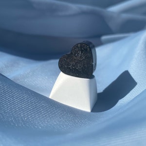 May include: A black heart-shaped ring with a sparkly finish, displayed on a white pedestal against a blue fabric background.
