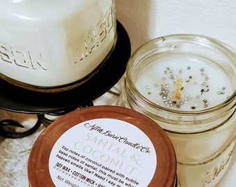 Puede incluir: Dos tarros de vidrio con tapas marrones llenos de cera de vela blanca. Los tarros están etiquetados con "After Burn Candle Co. Santal & Coconut" y tienen el texto "Top notes of coconut paired with subtle base notes of santal, this must be what heaven smells like? Need I say more? Soy Wax - Cotton Wick - Hand Poured Net Weight: 8 oz / 226 g".