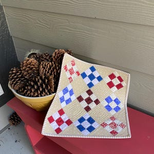 May include: A square quilted fabric mat featuring a nine-patch pattern with alternating cream-colored squares and smaller blocks of red, white, and blue fabric. The quilting displays a swirling stitch design throughout the cream background fabric.