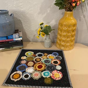 May include: A handmade, square fabric art piece with a black background and numerous colorful, circular fabric appliqués. The piece is bordered with a black and white striped fabric. Decorative vases and books are in the background.