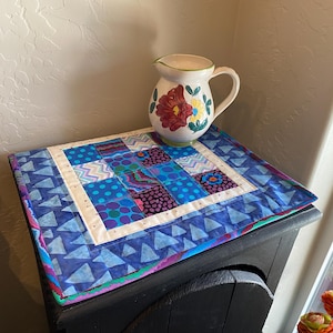May include: A handmade, square quilted placemat with a patchwork design in shades of blue, purple, and white. A white ceramic pitcher with a floral design in red, yellow, and green sits on top of the placemat. The pitcher has a curved handle and a spout.