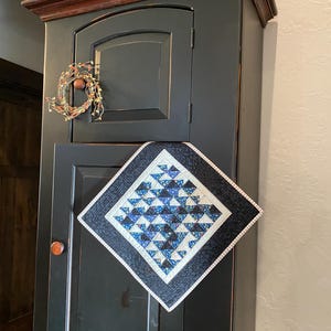 May include: A square quilted wall hanging featuring a geometric pattern of blue, black, and white triangles. The center design is framed by a thin white border and a wider black outer border with a subtle textured pattern, oriented diagonally.