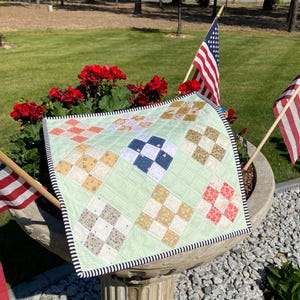 May include: A square quilted fabric piece featuring a light green background with a grid of nine-patch blocks in various floral and geometric patterns, finished with a black and white striped binding edge, displayed on a stone pedestal.