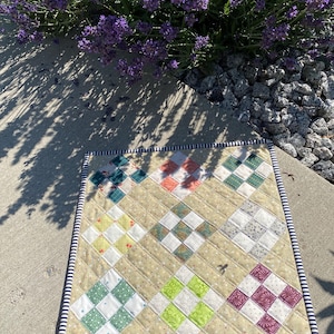 May include: A square fabric quilt featuring a nine-patch block pattern with various colored and patterned squares, including green, orange, yellow, and purple, set against a tan background with a navy and white striped border trim.