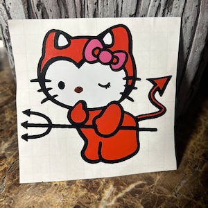 May include: A red and black cartoon illustration of Hello Kitty dressed as a devil holding a trident. She is wearing a red devil costume with a pink bow and has a winking eye.