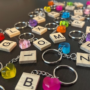 May include: A collection of colorful heart-shaped charms attached to keychains with letter tiles from a Scrabble game. The tiles spell out words like "GL", "OM", "I", "Z", "F", "K", "M", "L", "X", "P", and "B".