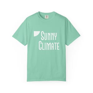 May include: A mint green t-shirt with the words "SUNNY CLIMATE" printed in white. The shirt has a crew neck and short sleeves. The text is in a stylized font with a white triangle on the left side.