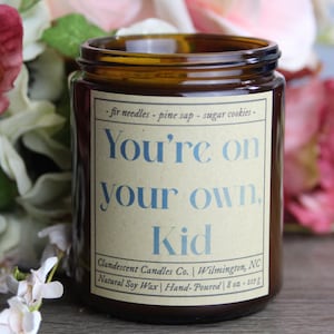 Op de afbeelding: Een bruine glazen kaars pot met een wit label waarop staat "You're on your own. Kid" en "Clandescent Candles Co. Wilmington, NC Natural Soy Wax Hand-Poured 8 oz - 227 g". De kaars pot is omringd door roze en witte bloemen.