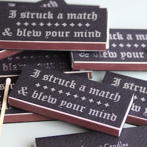 May include: A stack of black matchboxes with a white, gothic font design. The text on the matchboxes reads "I struck a match & blew your mind".