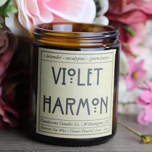 Puede incluir: Una vela en tarro de vidrio marrón con una etiqueta que dice "VIOLET HARMON". La etiqueta también enumera "lavanda - eucalipto - hojas verdes" y "cera de soja natural". La vela está rodeada de un ramo de flores.