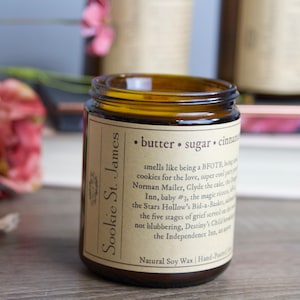 May include: A brown glass jar candle with a label that reads "Sookie St. James" and "butter. sugar. cinnamon". The label also includes a description of the scent, which is described as smelling like "being a BFOTR, being cookies for the love, super cool party, Norman Mailer, Clyde the cake, the Inn, baby #3, the magic rinse, the Stars Hollow's Bid-a-Basket, and the five stages of grief served on a blubbering, Destiny's Child, and the Independence Inn."