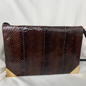 May include: A rectangular dark brown snakeskin clutch bag featuring a glossy finish and a textured scale pattern. The bag is accented with polished gold-tone metal corner protectors on the bottom left and right edges and includes a thin black strap.