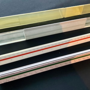 May include: Four architectural scale rulers with clear plastic cases. The rulers are white with red and green markings, and the cases are clear and yellowed. The scales are marked in centimetres.