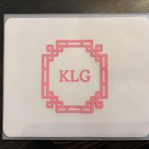 May include: A white card with a pink decorative frame and the letters "KLG" inside. The card is in a clear plastic sleeve. The background is a dark wood surface.