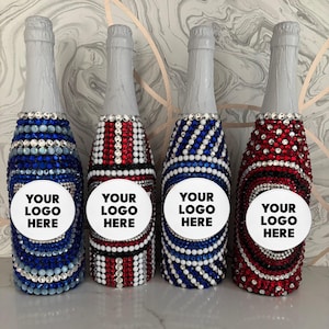 May include: Four champagne bottles decorated with colorful rhinestones. The bottles are adorned with blue, red, and white rhinestones, with a white circle that says "YOUR LOGO HERE". The bottles have silver foil tops.