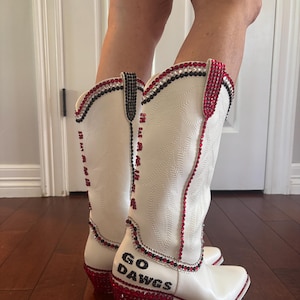 May include: White cowboy boots with red, black, and silver accents. The boots have a decorative pattern and the words "GO DAWGS" on the side. The heels and upper edges are embellished with red rhinestones.