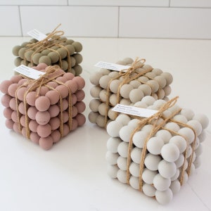 May include: Four sets of bubble coasters in various colors: dusty rose, sage green, light gray, and off-white. Each set is cube-shaped, made of small spheres, and tied with twine. Each set has a tag that reads "modernicki Bubble Coasters".