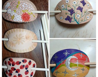 May include: A collection of five oval leather hair slides, each featuring a wooden stick. Designs include a sun and geometric patterns, white flowers with purple butterflies, embossed floral motifs, red ladybugs, and a silver moon with stars.