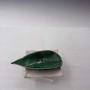 Puede incluir: Un jabonera de cerámica con forma de hoja verde con vetas detalladas. La hoja tiene tres orificios de drenaje y descansa sobre una base cuadrada de cerámica blanquecina. El diseño es sencillo y elegante.