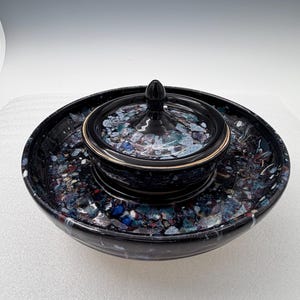 Puede incluir: Un juego de servir de cerámica negra con un patrón moteado y colorido. El juego incluye un cuenco grande, un cuenco más pequeño con tapa y un borde dorado. La tapa tiene un pequeño asa redondeada.