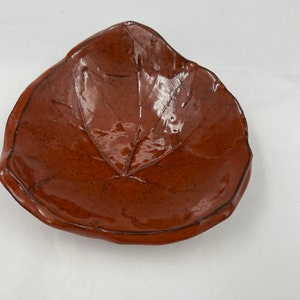 Puede incluir: Un plato de cerámica marrón con forma de hoja. El plato tiene un acabado brillante y un diseño de hoja en relieve.