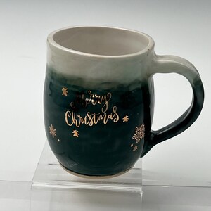 May include: A green ceramic mug with a white rim and a gold inscription that reads "Merry Christmas" with gold snowflake accents.