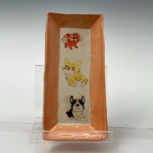 May include: A rectangular ceramic dish with a peach-colored rim and a white interior. The dish features three cartoon dogs in different poses and colors: a red dog, a yellow dog, and a black and white dog.