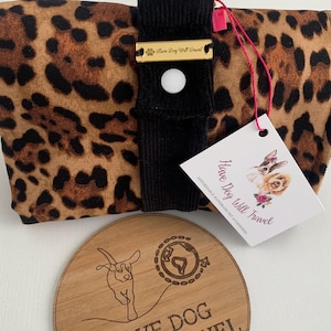 Puede incluir: Un transportín para perros de estampado de leopardo con una correa negra y una etiqueta dorada que dice "Have Dog Will Travel". El transportín está plegado y se encuentra sobre una superficie de madera con un letrero redondo de madera que dice "Have Dog Will Travel" con un dibujo de un perro corriendo con un globo terráqueo.