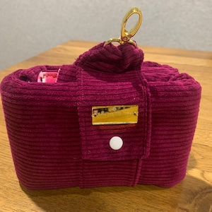 Könnte beinhalten: Eine bordeauxrote Cordtasche mit einem goldenen Reißverschluss und einem goldenen Etikett mit der Aufschrift "The Daily Edited".