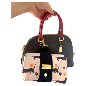 Puede incluir: Un bolso de mano marrón y negro con asas rojas y una cremallera dorada. Un bolso más pequeño con un diseño de dachshund y huellas de patas está sujeto al asa del bolso más grande. El bolso más pequeño tiene el texto "Happy Dog Will Travel".