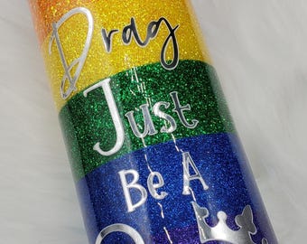 Puede incluir: Un vaso de glitter arcoíris con el texto "Don't Be A Drag Just Be A Queen" en letras plateadas. El vaso tiene un gráfico de corona en la parte inferior.