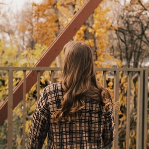 May include: A woman with long brown hair stands in front of a metal fence, looking out at a blurred background of fall foliage.