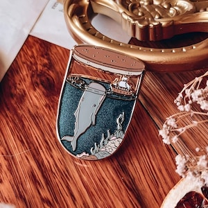 May include: A gold-toned enamel pin featuring a whale swimming in a glass jar with a small sailboat on the surface. The whale is grey and has a white belly. The jar is filled with a dark blue liquid and has a gold-toned lid.