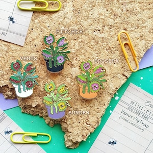 May include: Four enamel pins depicting different types of carnivorous plants. The pins are in the shape of Venus Fly Trap, Pitcher Plant, Sundew, and Drosera. Each pin has a gold outline and is set against a corkboard background.