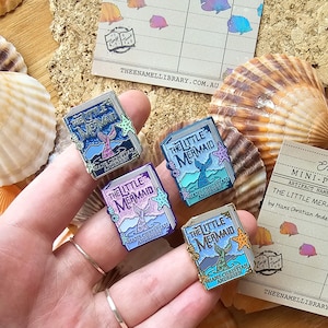 May include: Four enamel pins featuring a book cover design with the title "The Little Mermaid" by Hans Christian Andersen. The pins are in different colours, including blue, purple, and green, with a mermaid tail and a seashell design.