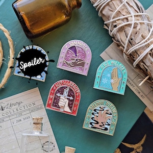 May include: A selection of enamel pins with fantasy themes. Each pin features a unique design and text, including "Summoner," "Ice Welder," "Metallurgist," and "Shadow Weaver." The pins are in various colours, with a black "Spoiler" pin.