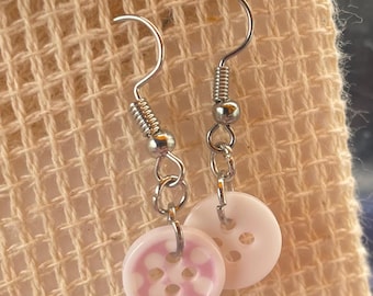 May include: A pair of handmade earrings featuring button-shaped charms. The earrings have silver-colored hooks and chains. One button is white with a pink floral design, and the other is a solid white button. The earrings are displayed on a beige fabric.