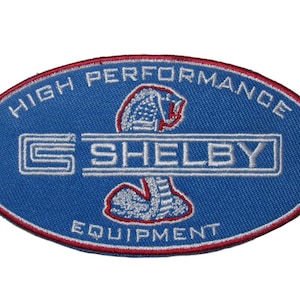 Op de afbeelding: Een blauwe ovale geborduurde patch met de woorden "High Performance Shelby Equipment" in witte letters rond een witte cobra met rode accenten.
