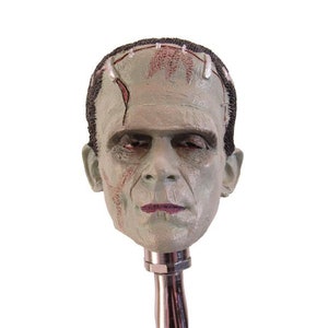 May include: A green and gray Frankenstein head bottle stopper with stitched scars on the forehead. The head is attached to a silver metal base.