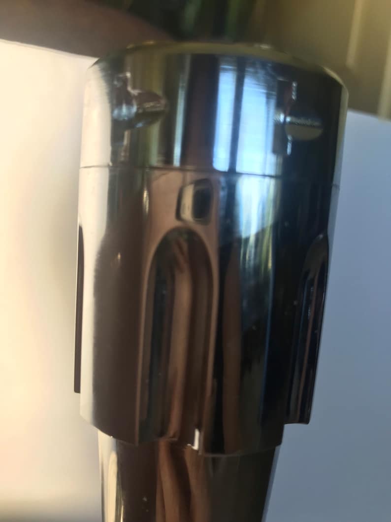 K&ouml;nnte beinhalten: Nahaufnahme eines verchromten Metall-Wasserhahn-Griffs mit strukturiertem Design. Der Griff hat zwei kleine Schrauben oben.
