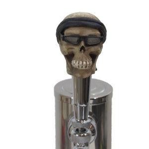 Puede incluir: Un tirador de cerveza con forma de cráneo con una bandana negra y gafas de sol. El tirador está hecho de un material marrón claro y tiene una base cromada.
