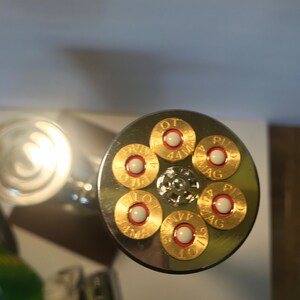 K&ouml;nnte beinhalten: Nahaufnahme eines silbernen Zylinders mit sechs goldfarbenen Patronenh&uuml;lsen. Jede H&uuml;lse hat eine wei&szlig;e Z&uuml;ndh&uuml;tchen und den Text "PILOT 44 MAG" aufgedruckt.