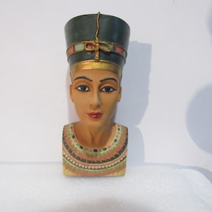 May include: A bust of an Egyptian queen wearing a blue and gold headdress with a pink and green band. The bust is made of painted resin and has a detailed necklace with pink, green, and black accents.